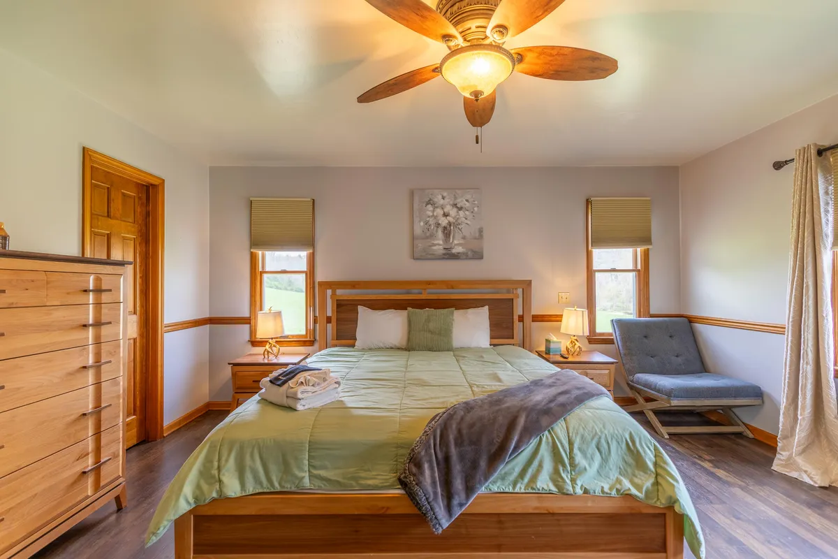 A serene king bedroom with green bedding, a wooden dresser, ceiling fan, and decorative artwork. Natural light streams through the window, creating a peaceful and inviting atmosphere perfect for relaxation.