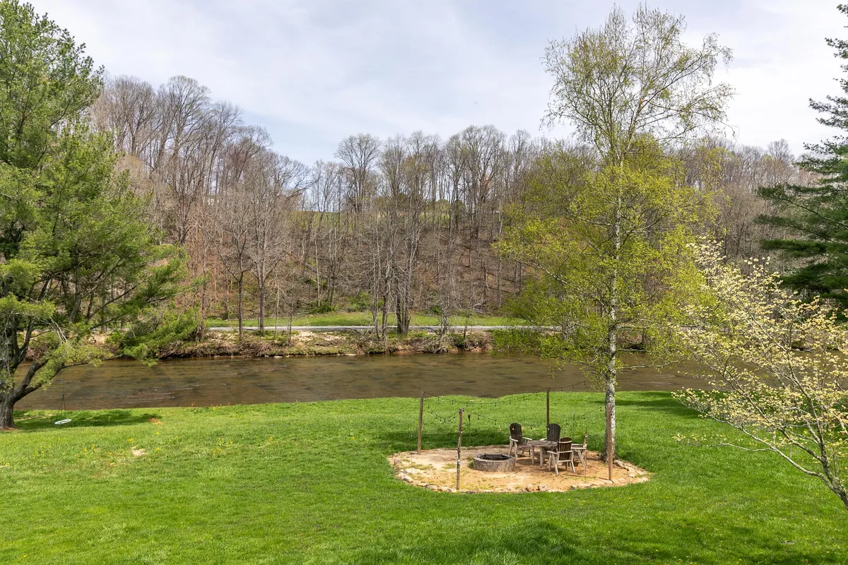 A wide view of the river bordered by a green lawn, with a cozy fire pit area set among the trees. The idyllic riverside scene beckons couples or friends to gather under the stars, roast s'mores, or savor morning coffee by the water.