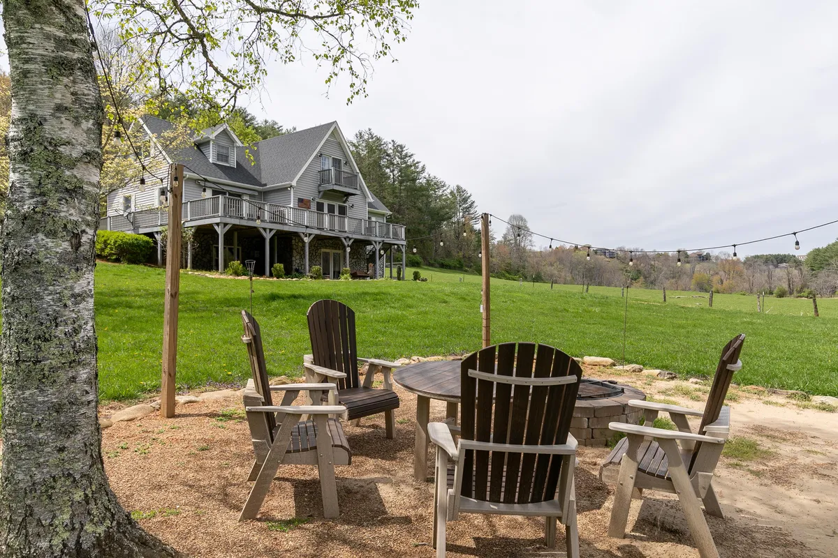 A welcoming fire pit area sits on a lush, green lawn with comfortable seating arranged for gatherings. The house is visible in the background, while the scene is surrounded by trees and open air, creating a relaxing, communal vibe. The fire pit invites stories and s’mores under sunset or starlit skies, perfectly facing the river and mountains beyond.