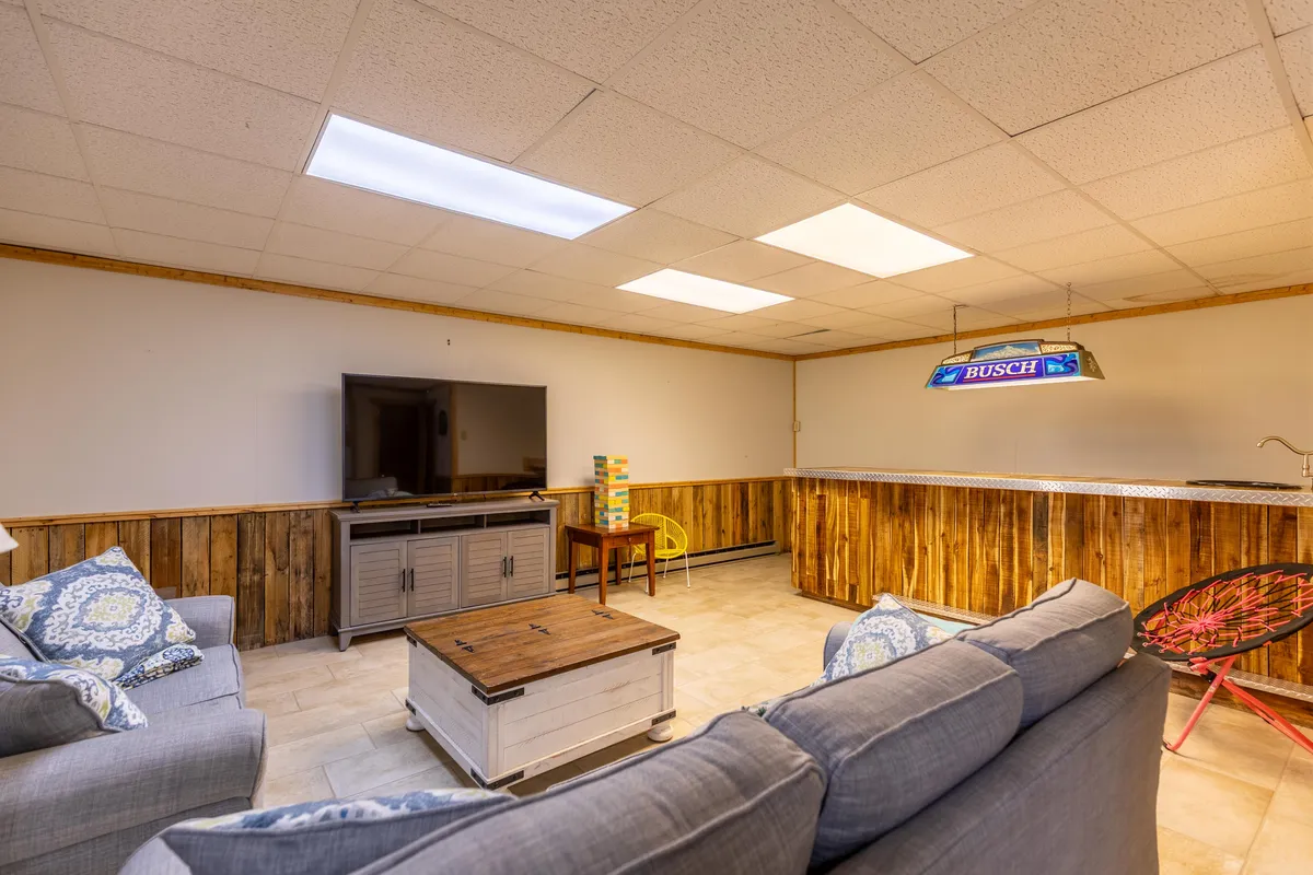 A relaxed lounge area with a plush sofa adorned with decorative cushions faces a coffee table and television, with a custom bar area visible in the background. The setup invites guests to unwind, watch movies, play games, or gather around the bar for drinks and conversation.