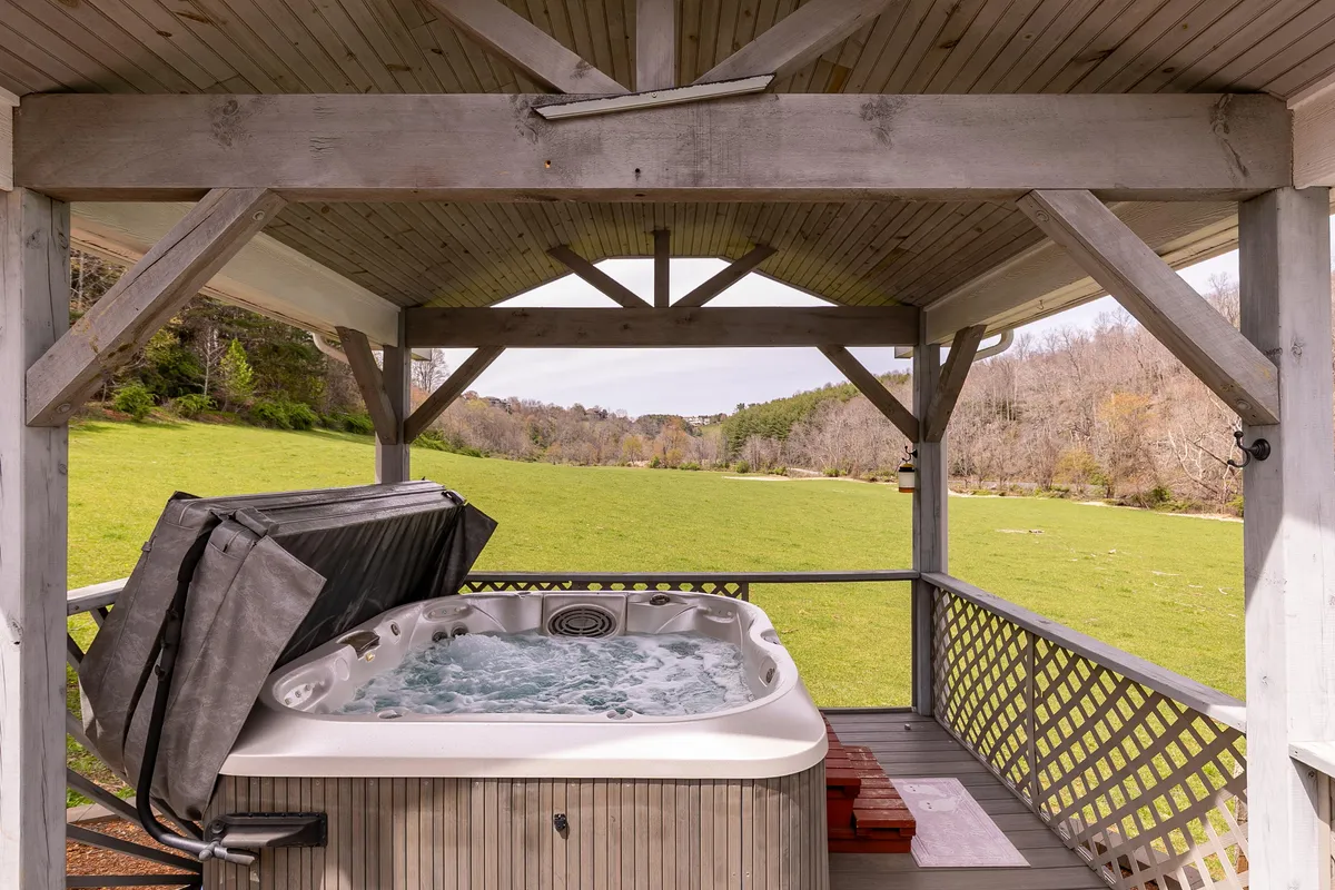 The image features a covered hot tub situated in an expansive green field next to a rustic wooden structure, perfect for soaking while enjoying open views or sunsets. The scene embodies relaxation with friends after a day of exploration.