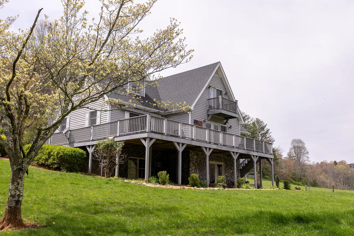 The house stands framed by a blooming tree, with the upper balcony clearly visible. People relax on the balcony and enjoy the manicured lawn below, perfect for gardening, picnics, or lawn games. The scene conveys a peaceful, lived-in atmosphere ideal for morning coffee or sunset appreciation.
