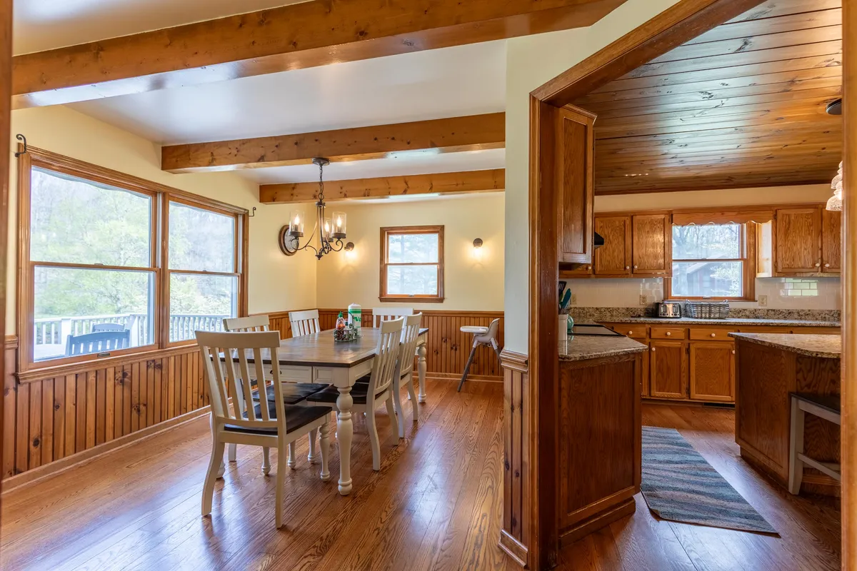 A spacious dining area with a large wooden table set beneath a chandelier. The dining space opens into the kitchen and is framed by large windows offering views of the surrounding landscape. The area is ideal for family meals, intimate dinners, and entertaining guests, capturing the spirit of shared occasions.