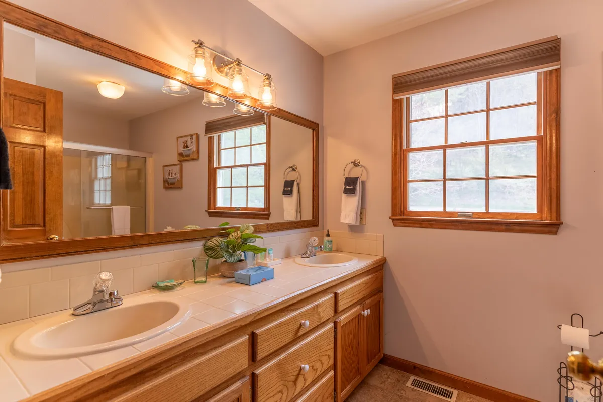 A spa-like bathroom with double sinks, large mirrors, wooden cabinetry, a window letting in natural light, and a decorative plant. The space feels calm, fresh, and perfect for self-care routines.