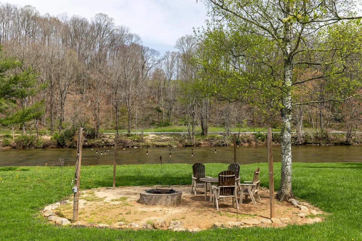 A fire pit surrounded by wooden chairs sits beside the river, embraced by trees and open sky. The setup suggests a relaxed environment for campfire gatherings and s’mores, with the natural river setting in clear view.