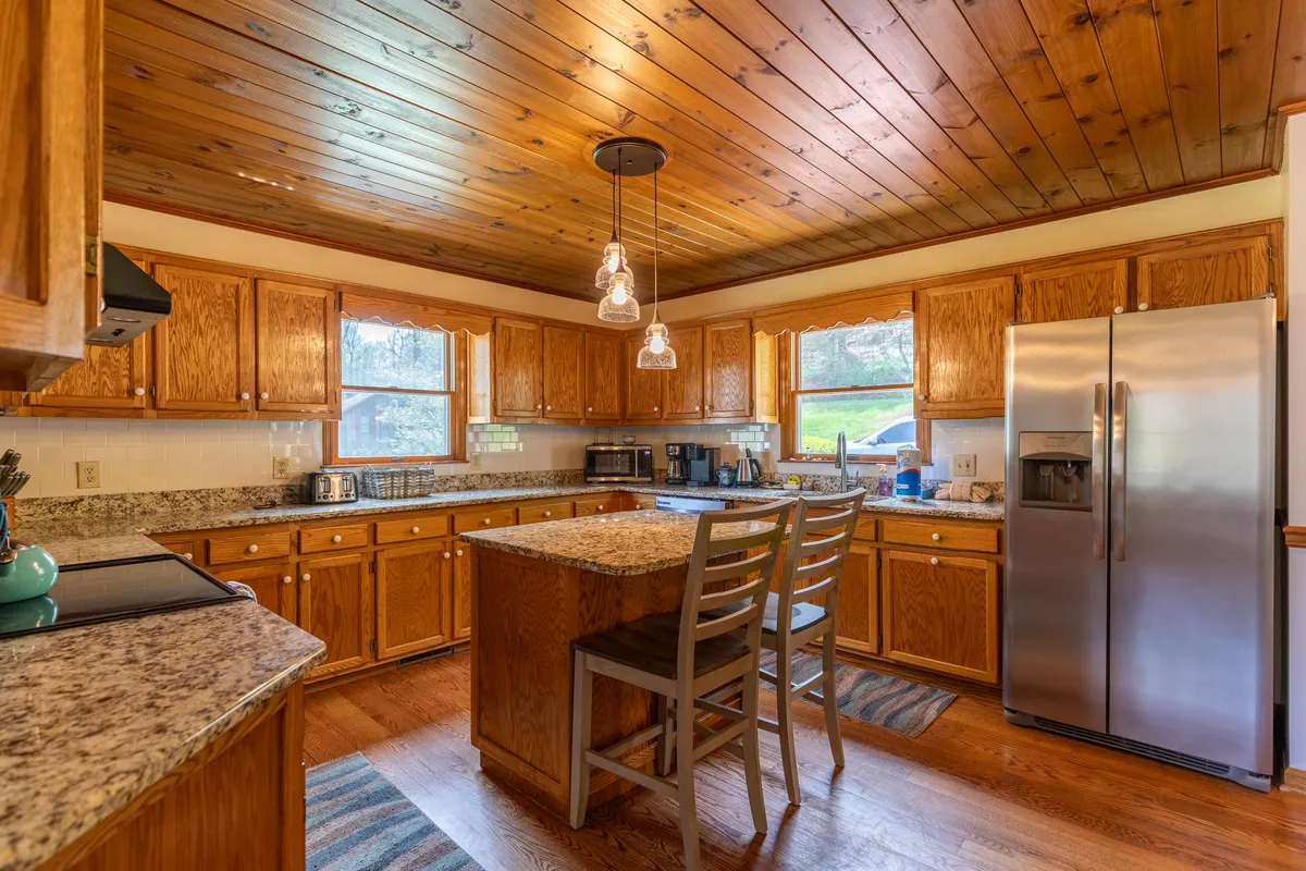 A modern, well-appointed kitchen featuring granite countertops, wooden cabinetry, an island with seating, and expansive windows. The space is designed for cooking, casual dining, and gathering, with ample natural light enhancing the welcoming atmosphere.