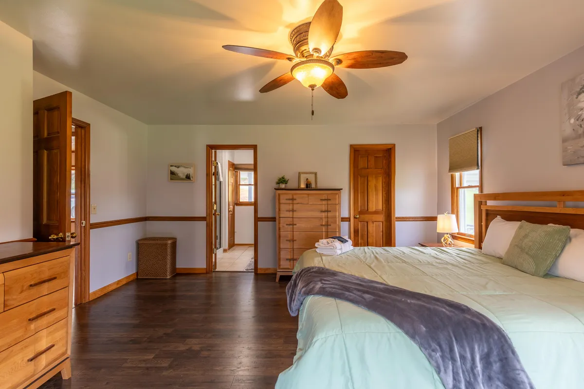 A calm and inviting king bedroom featuring soft bedding, a ceiling fan, a dresser, and doorways leading to other parts of the suite. The space is bathed in gentle light, reinforcing a soothing retreat ambiance.