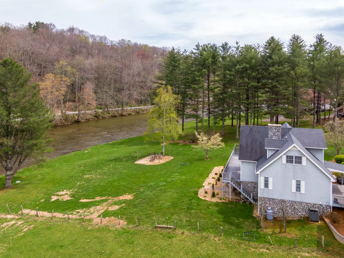 A peaceful exterior shot of Riverbank Retreat with the river meandering next to the house, mature trees, and an inviting outdoor seating area. The tranquil riverfront and greenery evoke a sense of privacy and connection with nature, while the seating area hints at relaxed gatherings or quiet moments.
