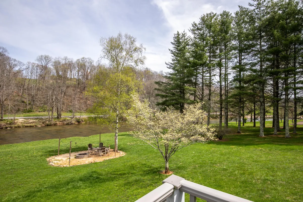 This exterior scene shows a peaceful riverside lawn with a fire pit surrounded by trees, creating an inviting space for gatherings, barbecues, and enjoying the outdoors at dusk or night.
