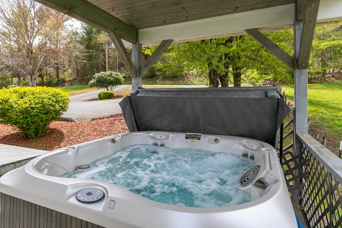A covered hot tub sits nestled on a wooden deck, surrounded by lush greenery and overlooking the river below. The inviting water and deck seating evoke relaxation and the opportunity to unwind in nature, whether in the morning with coffee or at night under the stars.