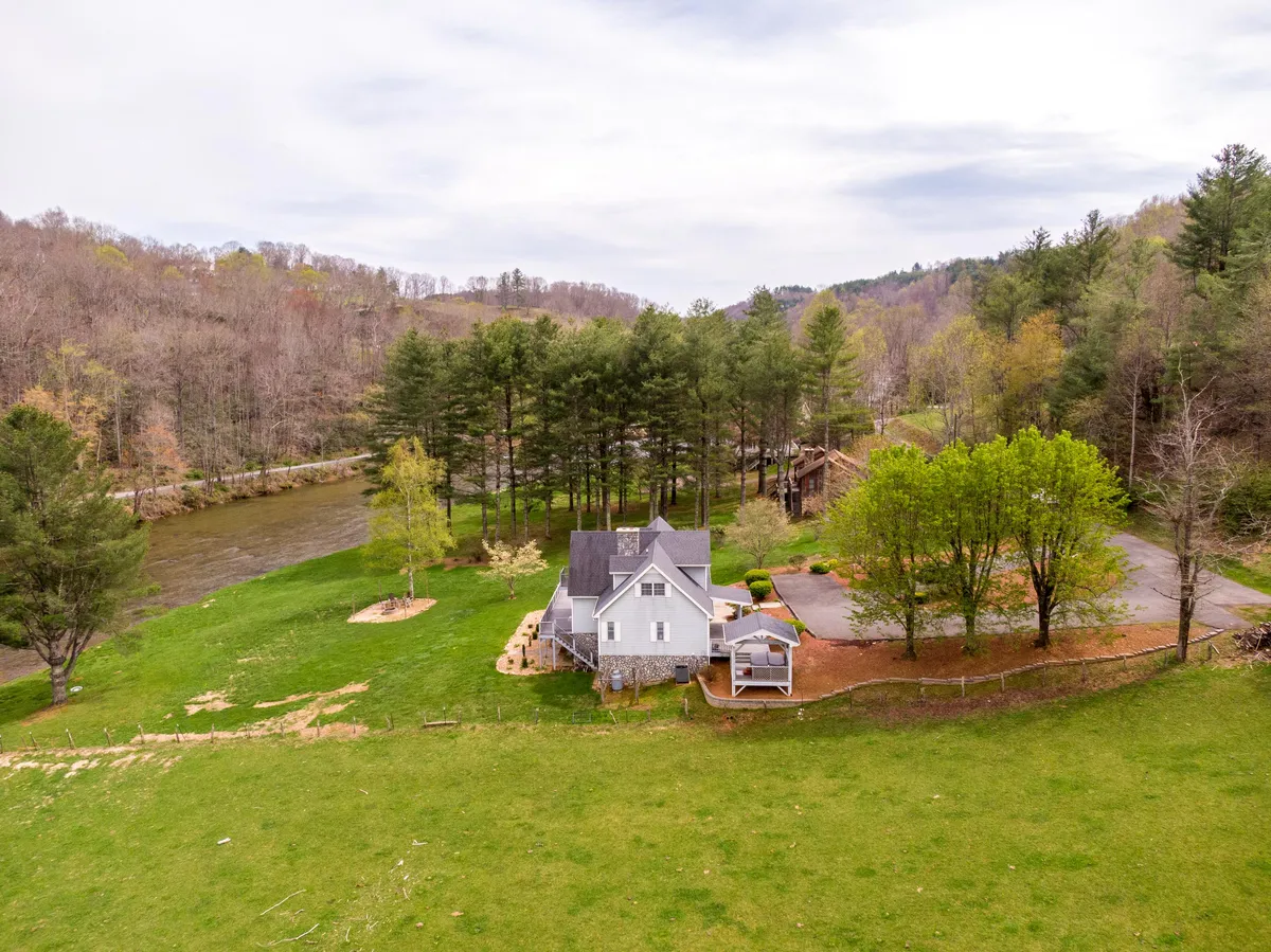 This image captures the house nestled beside the New River, with a sprawling green yard, mature trees, and the river running alongside the property. Children can be seen playing in the yard, and a campfire setup hints at evening family gatherings.
