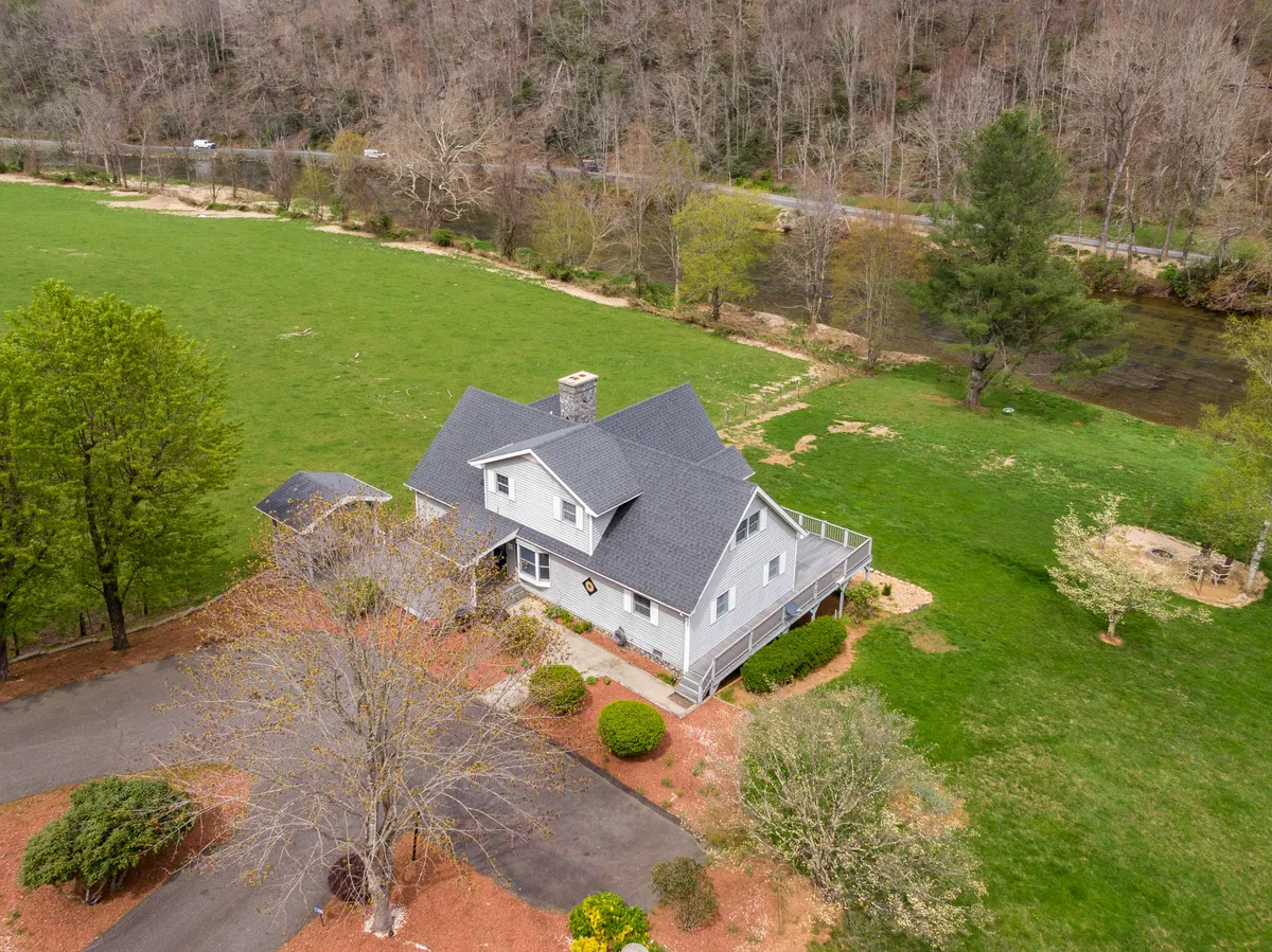 A broad exterior perspective featuring the house, lush greenery, the nearby river, and a welcoming driveway. The shot showcases the inviting porch and hints at the immersive natural landscape surrounding the retreat, capturing both home comforts and outdoor adventure potential.