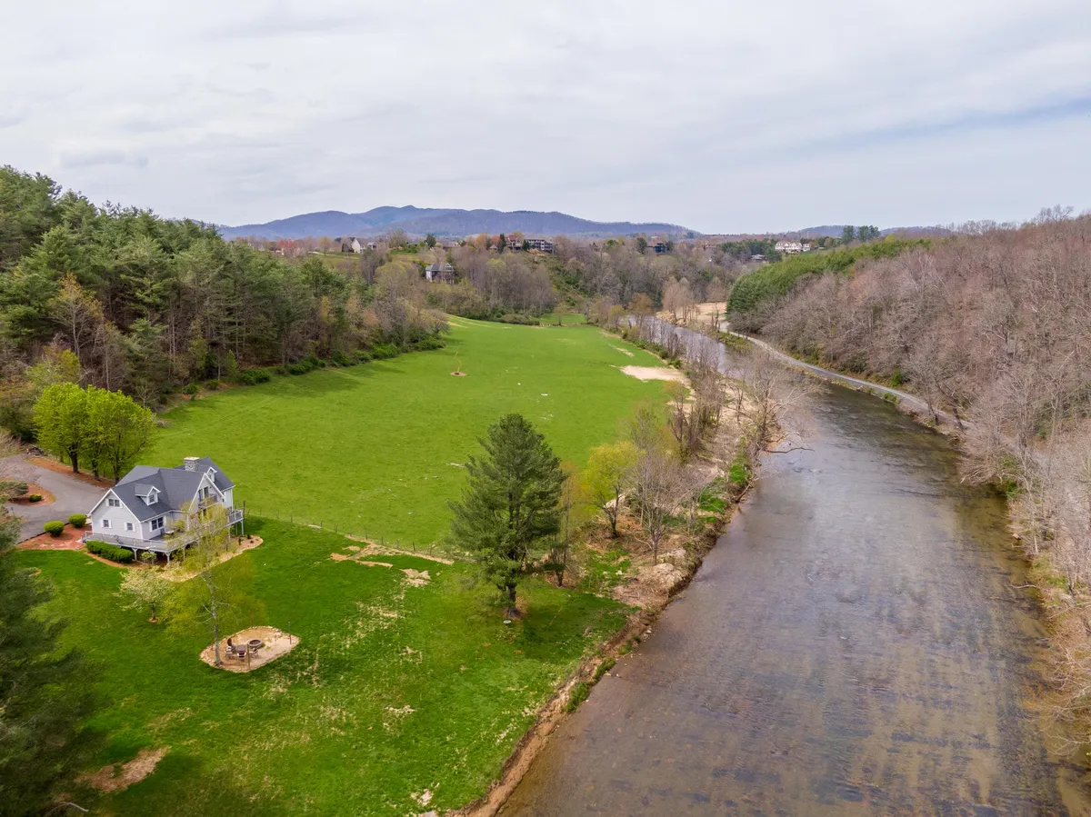 The image showcases a charming single-family mountain home nestled beside the tranquil New River, surrounded by lush green fields and mature trees. The serene river winds past the property, inviting outdoor activities like fishing, picnicking, and nature walks. As the sun sets, golden light washes over the landscape, creating an idyllic and peaceful atmosphere perfect for relaxation and wildlife observation.