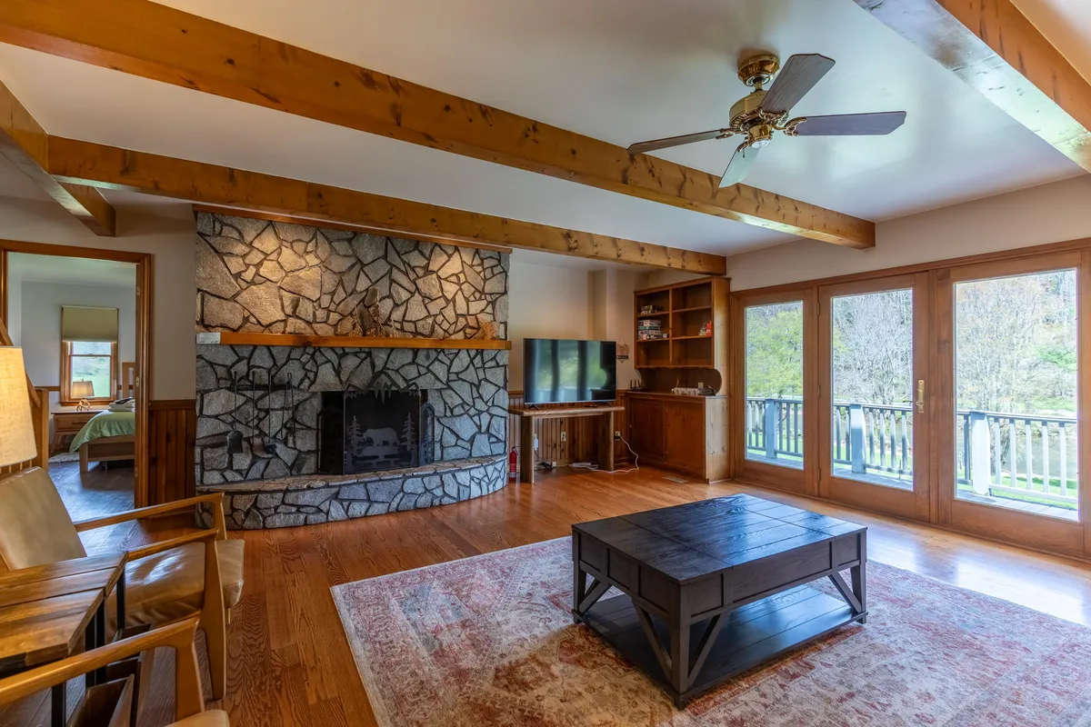 A warm, expansive living room dominated by a beautiful stone fireplace surrounded by wooden beams. Large windows allow natural light to flood the space, and a coffee table sits in front of a comfortable sofa, inviting guests to gather and relax. The room is arranged for family gatherings, reading, watching TV, and enjoying the views of nature.